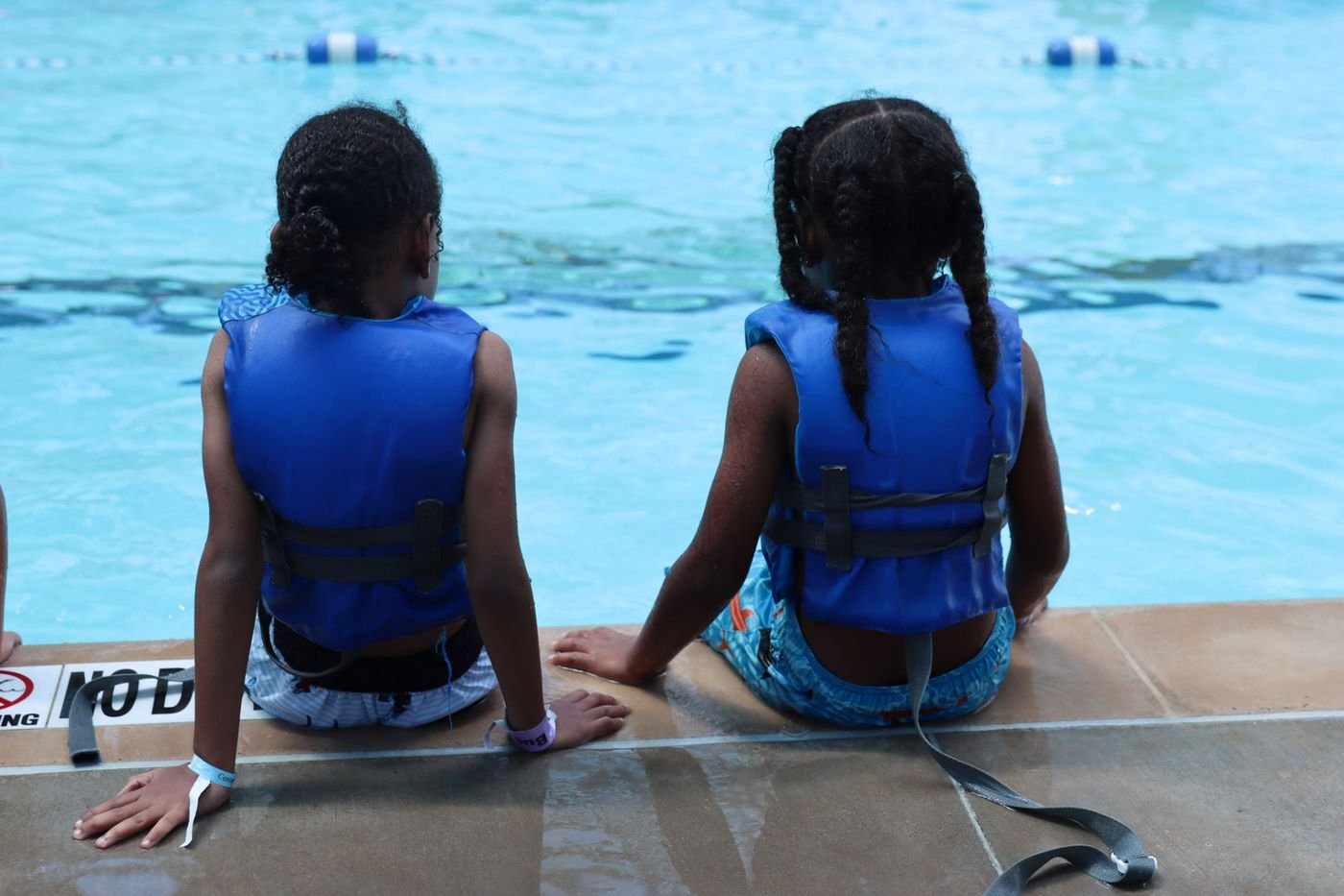 Two campers in life jackets sitting at the edge of a swimming pool